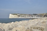A coastal landscape featuring a row of colorful houses along a beachfront beneath a large, white cliff. The sea is calm with gentle waves, and rocks are scattered in the foreground. The sky is mostly clear with a few scattered clouds.