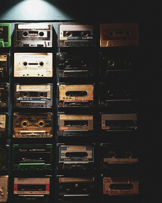 A collection of vintage audio cassette tapes arranged neatly in a grid pattern against a dark background. The tapes feature various designs and labels, showcasing a range of colors and types. A soft light illuminates the top portion, creating a nostalgic and retro vibe.