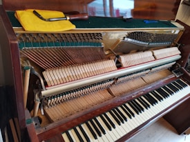 An upright piano with its top open, revealing the internal strings and hammers. A tuning lever and a yellow cloth are resting on top of the piano. The keys are visible, and the wood has a polished finish.