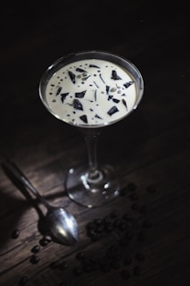 A glass filled with a creamy white dessert containing dark chocolate chunks and small spheres sits elegantly on a dark wooden surface. Scattered around the base of the glass are coffee beans, adding to the rich and sophisticated aesthetic. A silver spoon lies adjacent to the glass, suggesting readiness for indulgence.