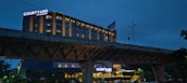 A hotel building with illuminated signs displaying the name 'Courtyard by Marriott'. The structure is well-lit, with multiple windows showing a warm glow against a dusky blue sky. An overhead bridge crosses in front of the hotel, and there are trees and streetlamps visible in the foreground.