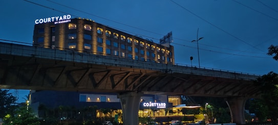A hotel building with illuminated signs displaying the name 'Courtyard by Marriott'. The structure is well-lit, with multiple windows showing a warm glow against a dusky blue sky. An overhead bridge crosses in front of the hotel, and there are trees and streetlamps visible in the foreground.