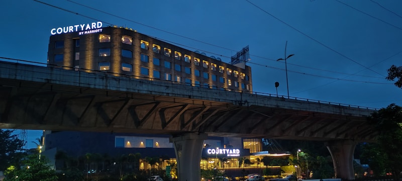 A hotel building with illuminated signs displaying the name 'Courtyard by Marriott'. The structure is well-lit, with multiple windows showing a warm glow against a dusky blue sky. An overhead bridge crosses in front of the hotel, and there are trees and streetlamps visible in the foreground.