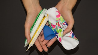 Two hands are holding several menstrual products including a blue menstrual cup, a white and red cloth pad, and two tampons wrapped in plastic.