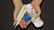 Two hands are holding several menstrual products including a blue menstrual cup, a white and red cloth pad, and two tampons wrapped in plastic.