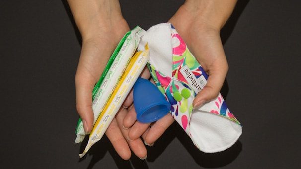 Two hands are holding several menstrual products including a blue menstrual cup, a white and red cloth pad, and two tampons wrapped in plastic.