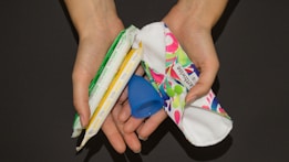 Two hands are holding several menstrual products including a blue menstrual cup, a white and red cloth pad, and two tampons wrapped in plastic.