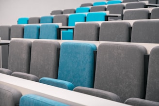A series of empty auditorium seats arranged in rows, featuring a combination of grey and cyan fabric upholstery. The seating is typical of a modern lecture hall or theater, designed for comfort and visibility.