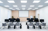 The room is a well-lit conference or training room with several rows of desks, each equipped with computer monitors and ergonomic chairs. A large projector screen is mounted on the beige wall, flanked by two plants. The room is illuminated by a grid of ceiling lights, and a whiteboard stands in the corner.