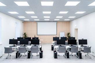The room is a well-lit conference or training room with several rows of desks, each equipped with computer monitors and ergonomic chairs. A large projector screen is mounted on the beige wall, flanked by two plants. The room is illuminated by a grid of ceiling lights, and a whiteboard stands in the corner.