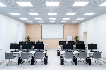 The room is a well-lit conference or training room with several rows of desks, each equipped with computer monitors and ergonomic chairs. A large projector screen is mounted on the beige wall, flanked by two plants. The room is illuminated by a grid of ceiling lights, and a whiteboard stands in the corner.