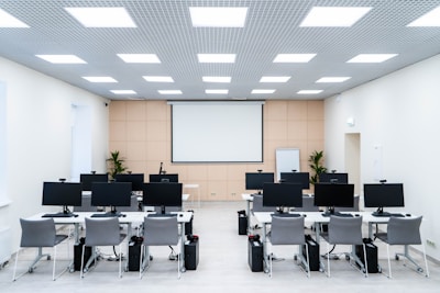 The room is a well-lit conference or training room with several rows of desks, each equipped with computer monitors and ergonomic chairs. A large projector screen is mounted on the beige wall, flanked by two plants. The room is illuminated by a grid of ceiling lights, and a whiteboard stands in the corner.