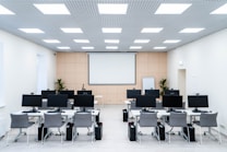 The room is a well-lit conference or training room with several rows of desks, each equipped with computer monitors and ergonomic chairs. A large projector screen is mounted on the beige wall, flanked by two plants. The room is illuminated by a grid of ceiling lights, and a whiteboard stands in the corner.