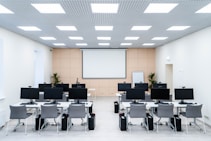 The room is a well-lit conference or training room with several rows of desks, each equipped with computer monitors and ergonomic chairs. A large projector screen is mounted on the beige wall, flanked by two plants. The room is illuminated by a grid of ceiling lights, and a whiteboard stands in the corner.