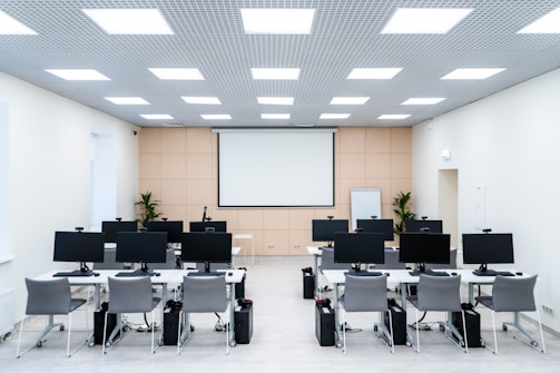 The room is a well-lit conference or training room with several rows of desks, each equipped with computer monitors and ergonomic chairs. A large projector screen is mounted on the beige wall, flanked by two plants. The room is illuminated by a grid of ceiling lights, and a whiteboard stands in the corner.