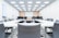 A modern, well-lit conference room with a large U-shaped arrangement of white tables and gray office chairs. The room features a ceiling with recessed lighting and a large screen or whiteboard at the front. Some computer monitors are visible toward the back, along with minimalistic decor and plants.