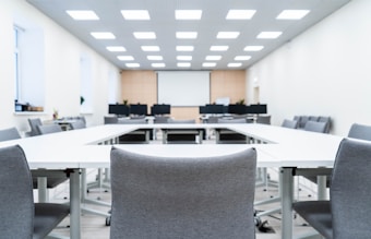 A modern, well-lit conference room with a large U-shaped arrangement of white tables and gray office chairs. The room features a ceiling with recessed lighting and a large screen or whiteboard at the front. Some computer monitors are visible toward the back, along with minimalistic decor and plants.