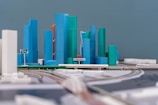 A detailed model of a modern city with high-rise buildings in blue and green shades. Some buildings have a white base, and an orange building is also noticeable. The structures are arranged along a curved road with a sign labeled 'D4'. A label in another language is visible in the foreground.