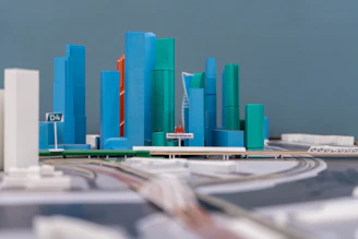 A detailed model of a modern city with high-rise buildings in blue and green shades. Some buildings have a white base, and an orange building is also noticeable. The structures are arranged along a curved road with a sign labeled 'D4'. A label in another language is visible in the foreground.