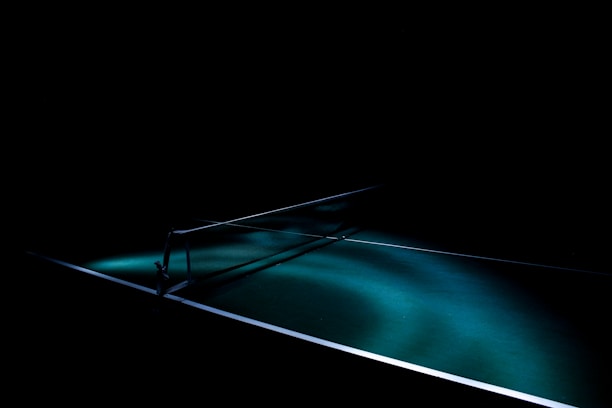 A dark setting with a spotlight illuminating a table tennis table, highlighting the net and a portion of the table surface. The lighting creates dramatic shadows and emphasizes the texture of the green table surface.