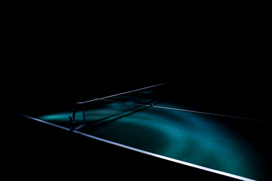 A dark setting with a spotlight illuminating a table tennis table, highlighting the net and a portion of the table surface. The lighting creates dramatic shadows and emphasizes the texture of the green table surface.