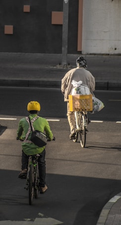 Two cyclists ride on a street, with one wearing a yellow helmet and green jacket, while the other carries a crate on the back of their bike. The road is marked with white lines, and the setting appears to be urban, possibly early morning or late afternoon based on the shadow angles.