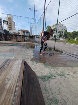 A person is cleaning a wet outdoor basketball or sports court with a broom. The court is enclosed by a tall fence, surrounded by urban buildings and some greenery.