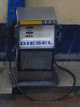 An old-fashioned diesel fuel pump located on a concrete slab. The pump has a metallic exterior with a hose attached, and the words 'DIESEL' and 'DED' prominently displayed. It's a rainy day, evident from the visible rain streaks and wet surfaces around the pump.