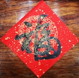 A decorative piece of art features intricate gold and black designs on a red square, possibly depicting stylized clouds and abstract Chinese characters. The artwork is set against a wooden background.