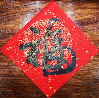 A decorative piece of art features intricate gold and black designs on a red square, possibly depicting stylized clouds and abstract Chinese characters. The artwork is set against a wooden background.