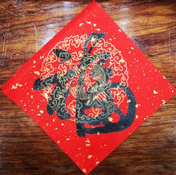 A decorative piece of art features intricate gold and black designs on a red square, possibly depicting stylized clouds and abstract Chinese characters. The artwork is set against a wooden background.