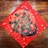 A decorative piece of art features intricate gold and black designs on a red square, possibly depicting stylized clouds and abstract Chinese characters. The artwork is set against a wooden background.