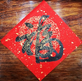 A decorative piece of art features intricate gold and black designs on a red square, possibly depicting stylized clouds and abstract Chinese characters. The artwork is set against a wooden background.