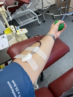 A close-up view of a person's arm during a blood donation. The arm is resting on a red cushioned chair and has a needle inserted, secured with adhesive tape. The person is holding a green stress ball, likely to aid in blood flow. Medical equipment and chairs are visible in the background.