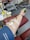 A close-up view of a person's arm during a blood donation. The arm is resting on a red cushioned chair and has a needle inserted, secured with adhesive tape. The person is holding a green stress ball, likely to aid in blood flow. Medical equipment and chairs are visible in the background.