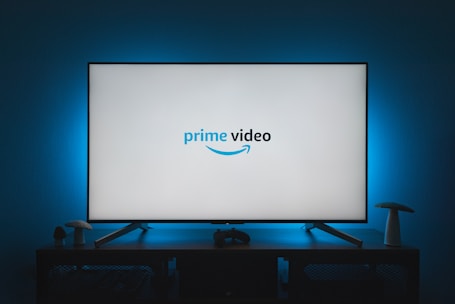A television screen displays the Prime Video logo, situated on an entertainment unit in a dimly lit room. The room is illuminated with a subtle blue backlight, creating a calming atmosphere. Two small decorative items, resembling mushrooms, are placed on either side of the unit.