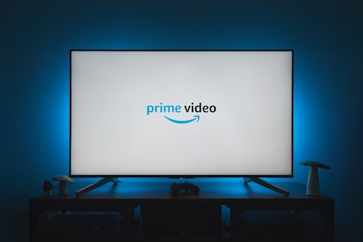 A television screen displays the Prime Video logo, situated on an entertainment unit in a dimly lit room. The room is illuminated with a subtle blue backlight, creating a calming atmosphere. Two small decorative items, resembling mushrooms, are placed on either side of the unit.