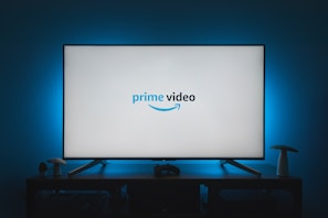 A television screen displays the Prime Video logo, situated on an entertainment unit in a dimly lit room. The room is illuminated with a subtle blue backlight, creating a calming atmosphere. Two small decorative items, resembling mushrooms, are placed on either side of the unit.