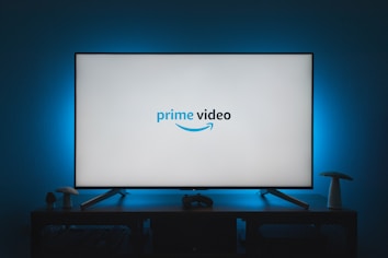 A television screen displays the Prime Video logo, situated on an entertainment unit in a dimly lit room. The room is illuminated with a subtle blue backlight, creating a calming atmosphere. Two small decorative items, resembling mushrooms, are placed on either side of the unit.