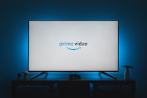 A television screen displays the Prime Video logo, situated on an entertainment unit in a dimly lit room. The room is illuminated with a subtle blue backlight, creating a calming atmosphere. Two small decorative items, resembling mushrooms, are placed on either side of the unit.
