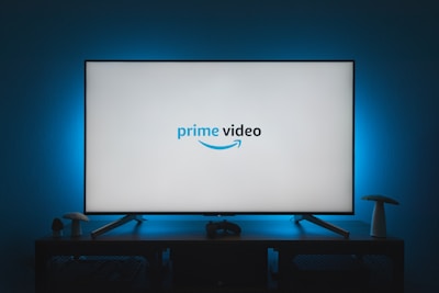 A television screen displays the Prime Video logo, situated on an entertainment unit in a dimly lit room. The room is illuminated with a subtle blue backlight, creating a calming atmosphere. Two small decorative items, resembling mushrooms, are placed on either side of the unit.