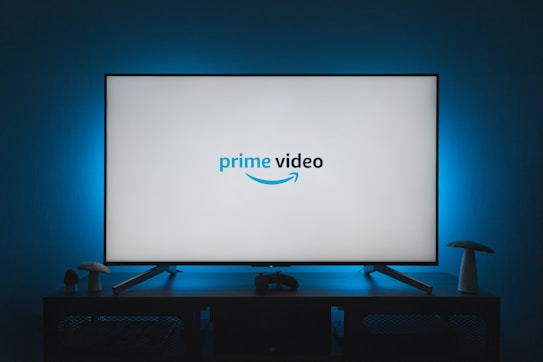 A television screen displays the Prime Video logo, situated on an entertainment unit in a dimly lit room. The room is illuminated with a subtle blue backlight, creating a calming atmosphere. Two small decorative items, resembling mushrooms, are placed on either side of the unit.