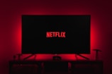 A dark room featuring a television screen displaying the word 'Netflix' in red against a black background. The screen is centered on a console table, with a video game controller placed in front. The room is dimly lit by red ambient lighting, creating a cozy atmosphere.