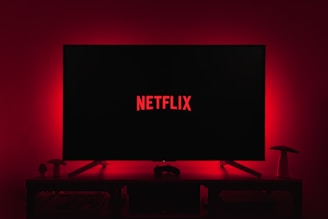 A dark room featuring a television screen displaying the word 'Netflix' in red against a black background. The screen is centered on a console table, with a video game controller placed in front. The room is dimly lit by red ambient lighting, creating a cozy atmosphere.