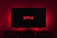 A dark room featuring a television screen displaying the word 'Netflix' in red against a black background. The screen is centered on a console table, with a video game controller placed in front. The room is dimly lit by red ambient lighting, creating a cozy atmosphere.