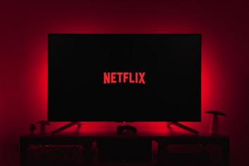 A dark room featuring a television screen displaying the word 'Netflix' in red against a black background. The screen is centered on a console table, with a video game controller placed in front. The room is dimly lit by red ambient lighting, creating a cozy atmosphere.