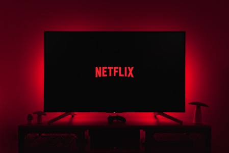 A dark room featuring a television screen displaying the word 'Netflix' in red against a black background. The screen is centered on a console table, with a video game controller placed in front. The room is dimly lit by red ambient lighting, creating a cozy atmosphere.