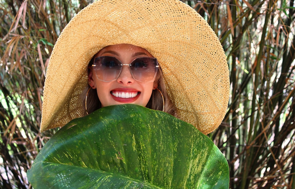 a woman in a straw hat and sunglasses holding a large green leaf