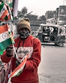 A person is holding several Indian flags in a street setting. They are wearing a red hoodie and a beige beanie, with a face mask covering their mouth. In the background, there is an auto-rickshaw with passengers and some buildings.