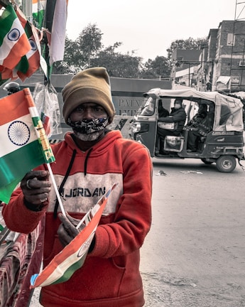 A person is holding several Indian flags in a street setting. They are wearing a red hoodie and a beige beanie, with a face mask covering their mouth. In the background, there is an auto-rickshaw with passengers and some buildings.